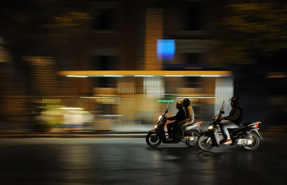 Two motorbike riders passing a lit shopfront at night
